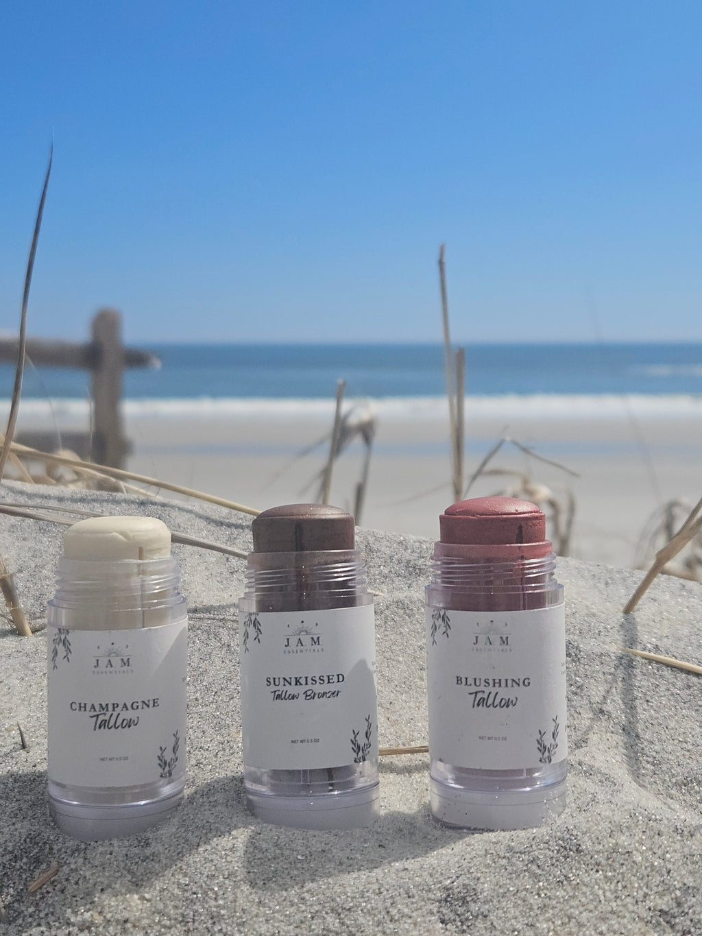Three JAM Beauty stick tallow products in clear plastic tubes standing upright in sand, with beach grass and water blurred in the background. The labels read Champagne Tallow, Sunkissed Tallow Bronze, and Blushing Tallow.