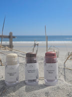 Three JAM Beauty stick tallow products in clear plastic tubes standing upright in sand, with beach grass and water blurred in the background. The labels read Champagne Tallow, Sunkissed Tallow Bronze, and Blushing Tallow.
