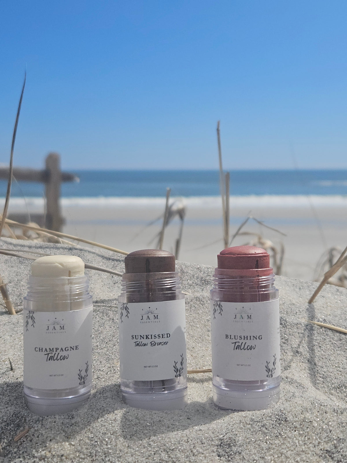 Three JAM Beauty stick tallow products in clear plastic tubes standing upright in sand, with beach grass and water blurred in the background. The labels read Champagne Tallow, Sunkissed Tallow Bronze, and Blushing Tallow.